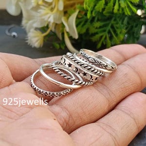 May include: A collection of silver rings with various designs, including stars, moons, and geometric patterns. The rings are stacked on a hand, showcasing their different textures and styles. The text "925jewells" is visible.