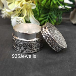May include: A round silver container with a removable lid featuring an intricate embossed floral pattern. The side of the base also displays a detailed engraved scrollwork design. The text 925Jewells is printed in white at the bottom left corner.
