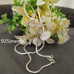 Puede incluir: Un collar de cadena de serpiente plateada con un diseño delicado. El collar se exhibe sobre un fondo de flores blancas y amarillas con follaje verde. El texto "925jewells" es visible en la esquina inferior izquierda.