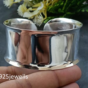 May include: A wide, polished silver cuff bracelet. The bracelet has a smooth, reflective surface and a slightly curved shape. The text "925jewells" is visible at the bottom of the image. The bracelet is a piece of jewellery.