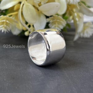 Può includere: Un anello largo in argento lucido dalla forma arrotondata. L'anello riflette la luce, creando un aspetto luminoso e brillante. L'anello è posto su uno sfondo scuro con fiori bianchi. Il testo "925Jewels" è visibile sul lato sinistro.
