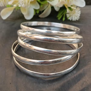 May include: Five silver bangles stacked together. The bangles are polished and reflective. The jewellery is presented on a dark surface, with white flowers in the background.