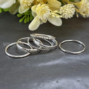 May include: A collection of silver rings in various designs. The rings include a plain band, a twisted rope design, a textured band, and a ring with a floral pattern. The rings are displayed on a dark surface with a blurred floral background.