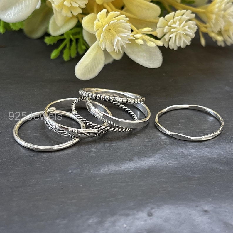 May include: A collection of silver rings in various designs. The rings include a plain band, a twisted rope design, a textured band, and a ring with a floral pattern. The rings are displayed on a dark surface with a blurred floral background.