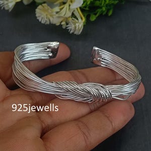 May include: Silver-toned cuff bracelet with a woven wire design. The bracelet features a knot-like center and open ends. The text "925jewells" is visible on the bracelet.