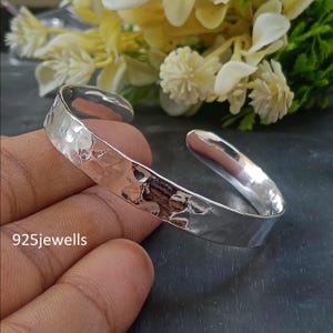 Puede incluir: Un brazalete de plata con textura martillada. El brazalete abierto se muestra sobre un fondo oscuro con flores blancas y amarillas. El texto "925jewells" es visible.