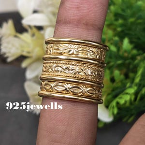 Può includere: Una pila di cinque anelli color oro indossati su un dito. Gli anelli presentano varie texture, tra cui fasce lisce, un motivo floreale inciso, un design a corda intrecciata e un motivo geometrico a forma di foglia. Il testo 925jewells è visibile.
