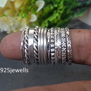 Puede incluir: Una colección de anillos de plata apilados en un dedo. Los anillos presentan varios diseños, incluyendo cuerdas trenzadas, bandas lisas y texturas estampadas. El fondo es borroso, con follaje verde y una superficie oscura. El texto "925jewells" es visible.