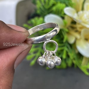 May include: A silver ring with a dangling charm featuring three small, round, silver beads. The ring is held up by a hand. The text "925Jewels" is visible on the image.