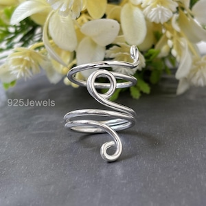 May include: A silver-colored ring with a spiral design. The ring wraps around the finger with a looped design at the top and a curled end. The text "925Jewels" is visible in the lower left corner. The background is a dark gray surface with white flowers.