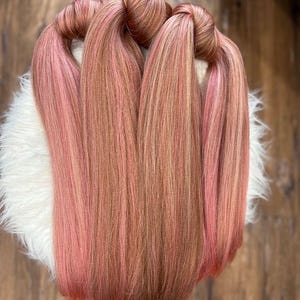 May include: Several bundles of hair extensions in shades of pink and brown are arranged on a white, fluffy surface. The hair is styled in long, straight strands, with the ends of the bundles knotted together.