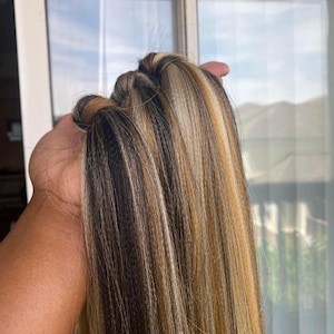 May include: A bundle of long, multi-toned hair extensions. The hair features a blend of black, brown, and blonde highlights, creating a natural and textured appearance. The extensions are held together at the top, showcasing the varied color strands.
