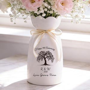 May include: A white ceramic vase with a bouquet of pink and white flowers, including roses and baby's breath. The vase has a scalloped top and a gold ribbon tied around the neck. Text on the vase reads "Happy 9th Anniversary" and "Love Grows Here."