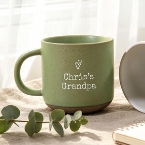 Puede incluir: Una taza de cerámica verde salvia con acabado moteado y base marrón oscuro, que presenta el texto blanco "Chris's Grandpa" y un pequeño icono de corazón, junto a una taza de cerámica blanca moteada con asa y base a juego.
