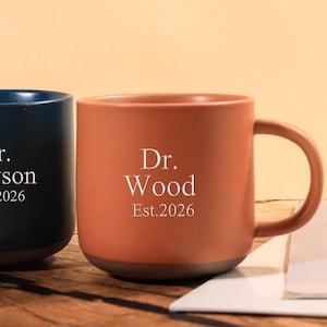 Puede incluir: Dos tazas de cerámica, una azul marino y otra terracota, están una al lado de la otra. Cada taza tiene un asa y presenta texto blanco: "Dr. Dawson Est.2026" y "Dr. Wood Est.2026". Las tazas están sobre una superficie de madera.