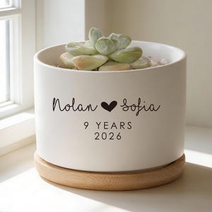 May include: A white cylindrical ceramic planter with a light wood base, containing a green succulent. The front features black text reading Nolan, a heart symbol, Sofia, 9 YEARS, and 2026, printed in a combination of script and sans-serif fonts.