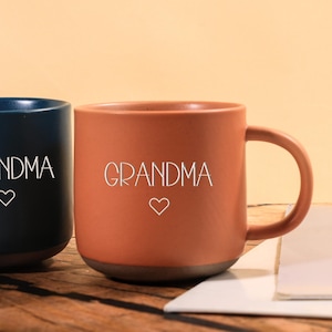 Puede incluir: Dos tazas de cerámica, una azul marino y otra terracota, se muestran sobre una superficie de madera. Cada taza tiene un asa y presenta la palabra "GRANDMA" en escritura blanca, con un pequeño símbolo de corazón debajo.