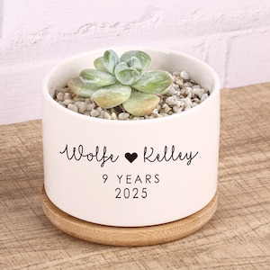 Puede incluir: Una maceta de cerámica blanca con una planta suculenta, pequeñas piedras y una base de madera. La maceta tiene el texto "Wolfe ♥ Kelley" y "9 YEARS 2025" en escritura negra. La maceta se encuentra sobre un posavasos redondo de madera marrón claro.