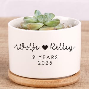 May include: A white ceramic plant pot with a succulent, on a wooden coaster. The pot is inscribed with the names "Wolfe" and "Kelly", a heart symbol, and the text "9 YEARS 2025".