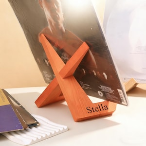 Pode incluir: Um suporte de discos de madeira em um tom laranja quente, projetado em forma de X. O suporte exibe um disco de vinil com a imagem de uma pessoa. A palavra "Stella" está gravada na base. Outros discos e um suporte de cerâmica branca também são visíveis.