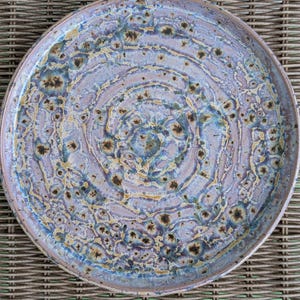 May include: A round ceramic plate with a unique, textured glaze. The plate features a spiral design in shades of lavender, blue, and gold, with dark brown speckles. The rim is a darker shade of lavender. This decorative plate is ideal for serving food.