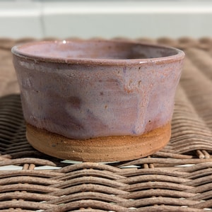 May include: A handmade ceramic bowl with a rustic aesthetic. The bowl features a light purple glaze over a reddish-brown base. The bowl's exterior has a textured surface, and the rim is slightly uneven.