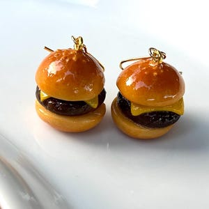May include: A pair of miniature hamburger earrings featuring a glossy brown bun, a dark brown patty, and a slice of yellow cheese. Each burger charm is attached to a gold-toned metal earring hook, creating a realistic food-themed accessory.