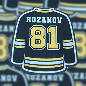 May include: A black and gold hockey jersey patch featuring the name "ROZANOV" above the number "81". The jersey has white and gold stripes on the sleeves and bottom. The background shows a blurred pattern of similar jersey patches.
