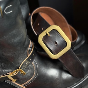 Puede incluir: Botas de cuero negro con hebillas y costuras doradas, combinadas con un cinturón de cuero marrón oscuro con una hebilla grande, cuadrada y dorada. Las botas tienen un diseño clásico y robusto.