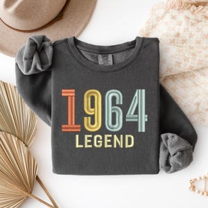 May include: Dark gray sweatshirt with the year "1964" embroidered in orange, yellow, and teal, with the word "LEGEND" below. The sweatshirt is folded, with a tan hat and decorative items in the background.