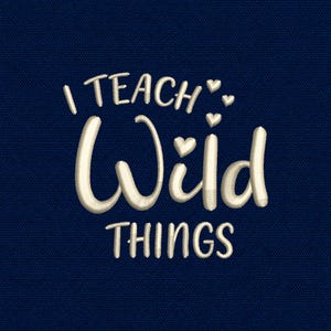 May include: An embroidery design on a dark blue fabric background featuring the white text I TEACH WILD THINGS in a mix of block and script fonts. The design includes three small white embroidered hearts positioned near the word Wild.