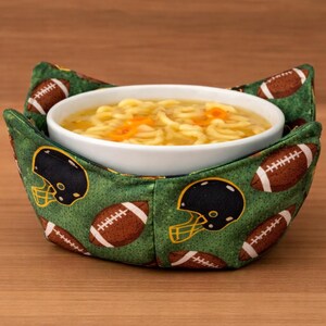 May include: A white ceramic bowl filled with soup sits inside a green fabric bowl cozy. The cozy is decorated with brown footballs and black and yellow football helmets. The soup has yellow noodles and orange carrots.