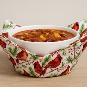 May include: A white ceramic bowl filled with chili, set in a fabric bowl cosy. The cosy has a white background with a pattern of red cardinals, green pine needles, and red berries. The chili contains beans and corn.