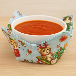 以下が含まれることがあります： 白いセラミックボウルに入ったトマトスープが、水色の布製のボウルホルダーに置かれています。ボウルホルダーには、子猫、キャンディケイン、クリスマスツリーをあしらったクリスマスをテーマにしたプリントが施されています。ボウルホルダーはボウルを保持するように設計されています。