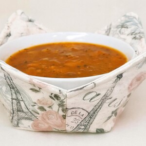 May include: A white ceramic bowl filled with orange soup, resting in a decorative bowl cozy. The cozy features a Parisian theme with Eiffel Tower and floral designs. The text "DE LA 25" is visible on the cozy.