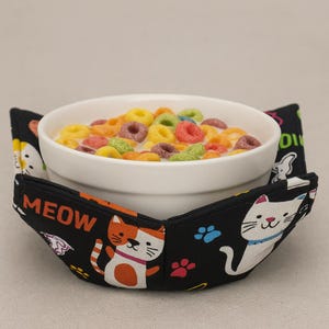 以下が含まれることがあります： カラフルなシリアルとミルクが入った白いセラミックボウルが、黒いボウルカバーの上に置かれています。カバーには猫のイラスト、足跡、そして「MEOW」という文字が描かれています。