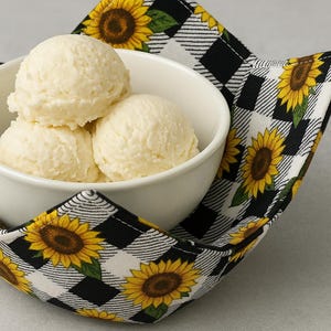 May include: A white bowl filled with three scoops of vanilla ice cream, resting on a black and white checkered bowl cozy. The cozy is decorated with yellow sunflowers.