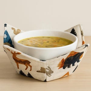 Op de afbeelding: Een witte keramische kom gevuld met soep rust in een crèmekleurige stoffen komhouder. De komhouder heeft een bosdierenprint met herten, beren, konijnen en bomen. De soep lijkt een groentesoep te zijn.