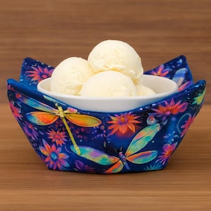 Puede incluir: Un cuenco blanco lleno de helado de vainilla se encuentra dentro de un cuenco de tela azul. El cuenco de tela está decorado con libélulas y flores de colores. El fondo es una superficie de madera.