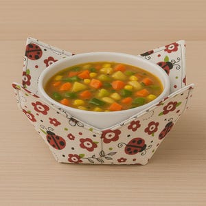 May include: A white ceramic bowl filled with vegetable soup, resting in a fabric bowl cozy. The cozy is cream-colored and decorated with red ladybugs and flowers. The soup contains carrots, corn, potatoes, and green beans.