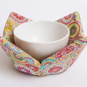 May include: A white ceramic bowl nestled in a colorful fabric bowl cozy. The cozy features a vibrant paisley pattern with red, yellow, blue, and green hues. The bowl and cozy are set against a white background.