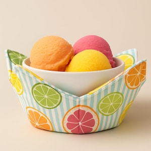 May include: A white bowl filled with scoops of orange, pink, and yellow ice cream, nestled in a decorative fabric bowl. The fabric bowl features a light blue and white striped pattern with colorful citrus fruit designs, including oranges, limes, and lemons.