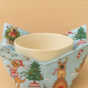 Puede incluir: Un protector de cuenco azul claro con un cuenco color crema en el interior. El protector presenta un patrón temático navideño con perros, árboles de Navidad y bastones de caramelo. El protector está diseñado para sostener un cuenco de comida.