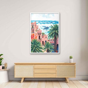 May include: A framed art print depicts a coastal scene with pink buildings, palm trees, and a blue ocean. The artwork features a white frame and is displayed on a light-colored wooden cabinet. The painting showcases a vibrant, sunny day.