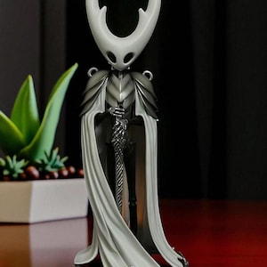 May include: A detailed figurine of a character with a white, curved headpiece and a flowing white cape. The figure is clad in silver armor and holds a detailed sword. The background is dark, with a plant in a white pot visible on the left.