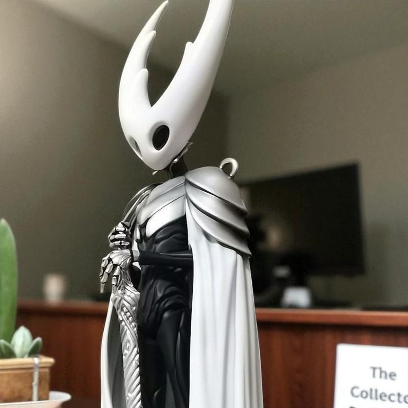 May include: A detailed figurine of a character from the video game Hollow Knight. The figure features a white head with two horns, silver armor, a white cape, and a silver sword. The character is posed in a standing position.