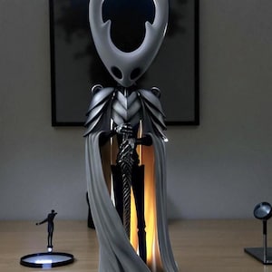 May include: A detailed figurine of a character from the video game Hollow Knight, featuring a white head and cape, silver armor, and a black sword. The figure is illuminated from within, creating a glowing effect. A small figure stands on a circular base.