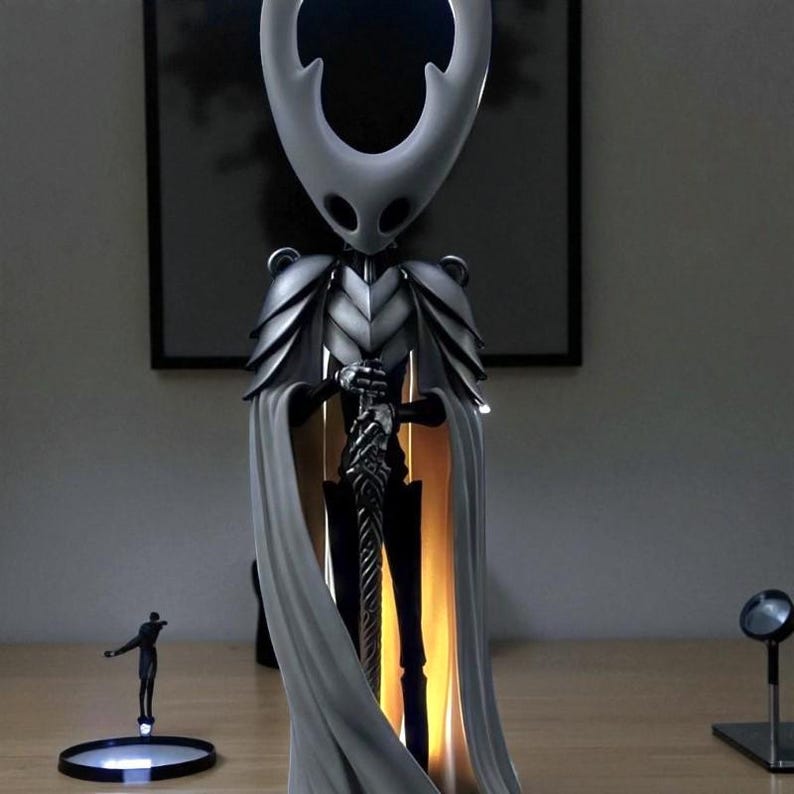 May include: A detailed figurine of a character from the video game Hollow Knight, featuring a white head and cape, silver armor, and a black sword. The figure is illuminated from within, creating a glowing effect. A small figure stands on a circular base.