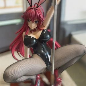 May include: A figurine of a woman in a black bunny suit, fishnet stockings and red high heels. The figure has long, red hair and bunny ears. She is holding a silver pole.