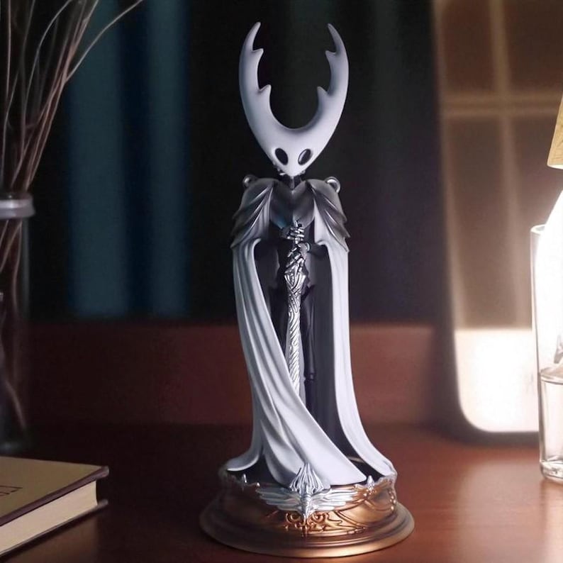 May include: A detailed figurine of a character with a white head and horns, wearing a dark cloak and holding a sword. The figure stands on a gold-colored base with intricate designs. The overall design is detailed and artistic.